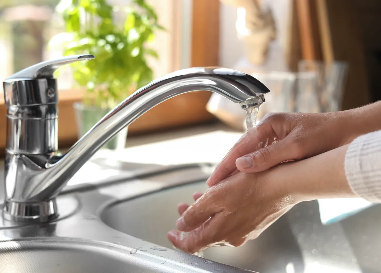 Person wäscht sich die Hände unter einem laufenden Wasserhahn in einer Küche.